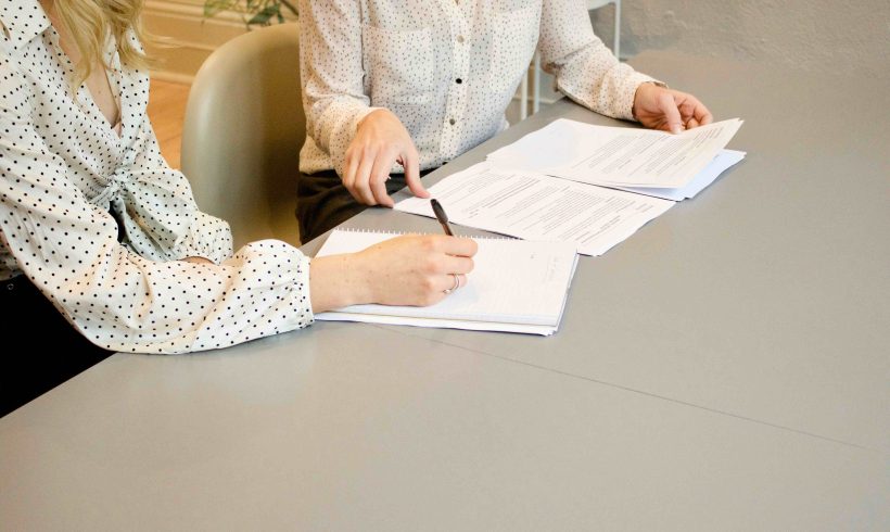 On aperçoit deux femmes qui échangent assises autour d’une table en lisant des documents contractuels. L’une tient un stylo pour prendre des notes et l’autre prend connaissance du contenu des documents.